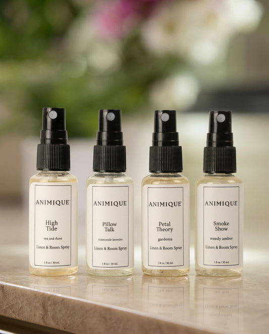 Four sample size bottles of room and linen spray by Animique — pet-safe home fragrance on a table with a dog sitting in the background.