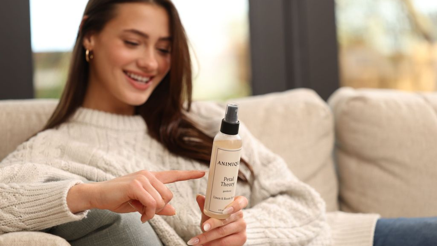 Woman sitting on a couch holding a bottle of Petal Theory room and linen spray by Animique — pet-safe home fragrance reading the ingredients