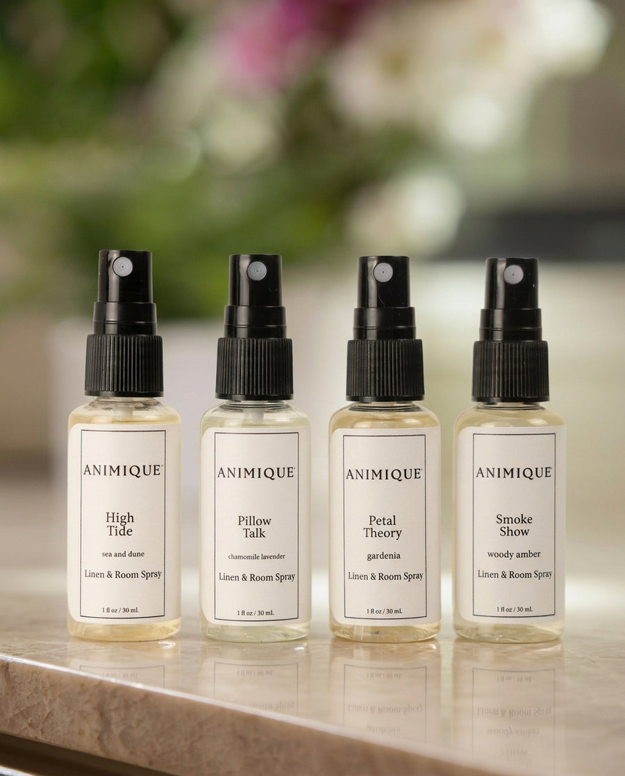 Four sample size bottles of room and linen spray by Animique — pet-safe home fragrance on a table with a dog sitting in the background.